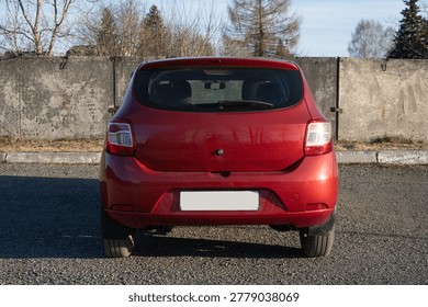 A rear view of a compact red hatchback parked on a gravel lot, showcasing its sleek design against a backdrop of a rustic wall and trees.