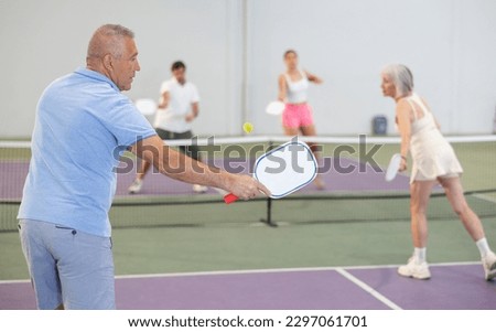 Rear view of concentrated sporty elderly man playing pickleball on indoor court, swinging paddle ready to return ball..