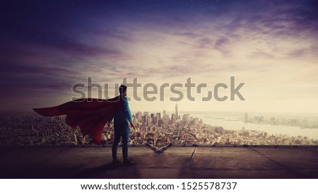Rear view confident businessman, red cape suit, as hero stands on the rooftop of a skyscraper looking over the city horizon. Superhero leadership and success concept. Surreal super power metaphor.