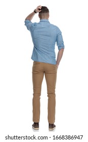 Rear view of confused casual man scratching his head while wearing blue shirt, standing on white studio background