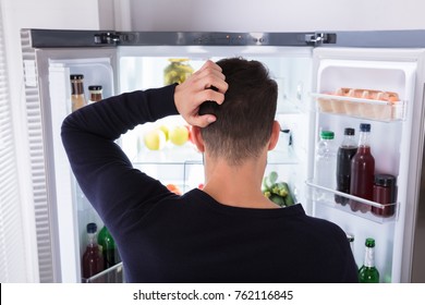 Rear View Of A Confused Young Man Looking At Food In Refrigerator