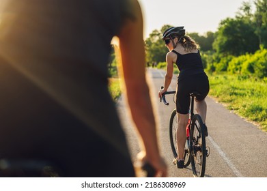 Rear view of a couple of athletes dressed in active wear biking fast on paved road. Countryside area, sunny day outdoors. Focus on a woman looking at her back. Concept of endurance and strength. - Powered by Shutterstock