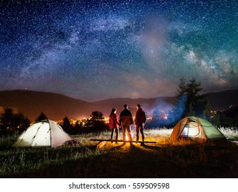 Rear View Couple Hikers Holding Hands And Their Friend Stands Near Campfire And Looks At It Under Night Sky With Stars On The Background Mountains And Luminous Town