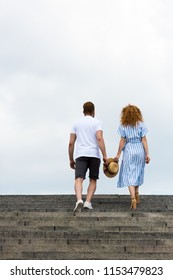 rear view of couple holding straw hat and walking on stairs against cloudy sky