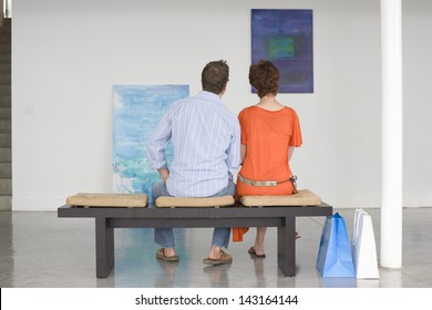 Rear View Of Couple Looking At Paintings While Sitting On Bench In Art Gallery