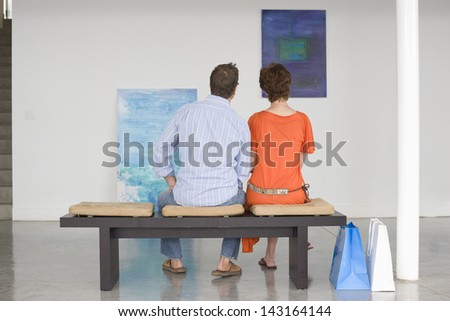 Rear view of couple looking at paintings while sitting on bench in art gallery