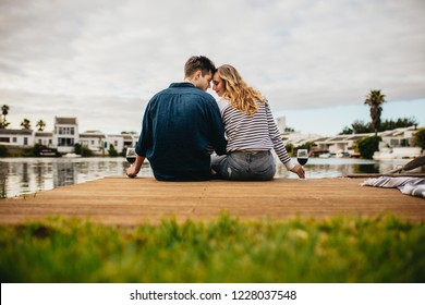 Rear View Of A Couple In Love Sitting Together Touching Their Heads Near A Lake. Couple On A Day Out Sitting Together Holding Wine Glasses On The Wooden Dock Near A Lake.