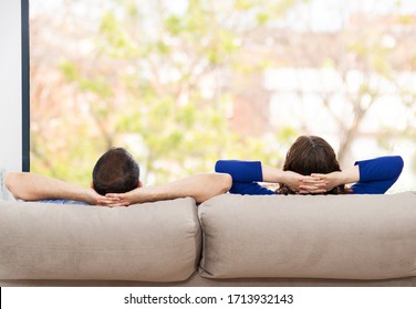 Rear View Of A Couple Relaxing On A Sofa At Home And Looking Outside A Green Background Through The Window Of The Living Room