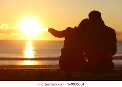 Rear view of a couple silhouette sitting cuddling and enjoying pointing at sun at sunset outside on the beach in winter