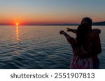 Rear view of a couple silhouette standing cuddling and enjoying pointing at the sun at sunset outside on the beach