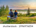 Rear view of couple sitting on hill top against beautiful summer landscape with forest and lake in summer evening at sunset. Tourists man and woman admiring wonderful northern nature. Vacation concept