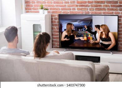 Rear View Of Couple Sitting On Couch Watching Television At Home