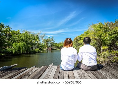 Rear View Of Couple Sitting On Wooden Bridge Over Lake