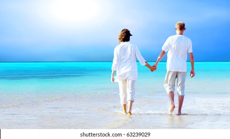 Rear View Of A Couple Walking On The Beach, Holding Hands.