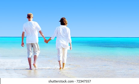 Rear View Of A Couple Walking On The Beach, Holding Hands.