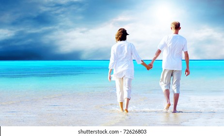 Rear View Of A Couple Walking On The Beach, Holding Hands.