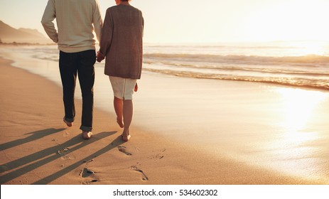 Rear view cropped shot of a senior couple holding hands walking on the beach. Mature couple together taking a stroll on the sea shore at sunset.