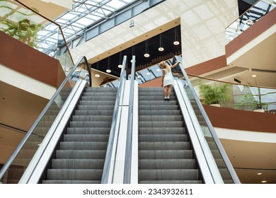 Rear view of cute blonde little girl 5-6 year old in white dress climbs escalator upstairs. Shot of adorable funny kid going up, back view from behind. Happy joy childhood concept. Copy ad text space