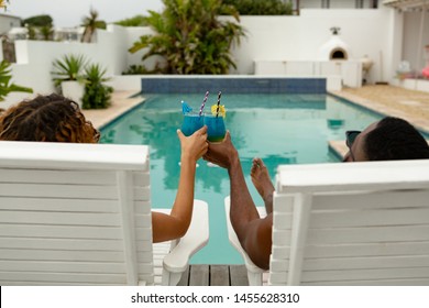 Rear View Of Cute Diverse Couple Toasting Glasses Of Cocktail While Relaxing On A Sun Lounger Near Swimming Pool In The Backyard. Summer Fun At Home By The Swimming Pool