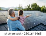 Rear view of dad with girl on roof with solar panels, hugging. Rooftop solar or photovoltaic system. Sustainable future for next generation.