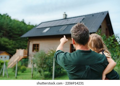 Vista trasera de papá sosteniendo a su niñita en brazos y mostrando en su casa con paneles solares instalados. Energía alternativa, ahorro de recursos y concepto de estilo de vida sostenible.