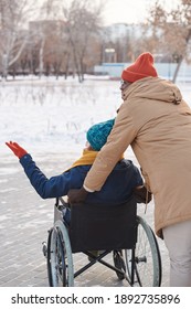 Rear View Of Disabled Woman In Wheelchair Walking In Winter Day With Assistant