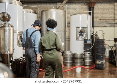 Rear view of diverse team of two cider factory workers inspecting production process on workshop floor with huge fermentation tanks at plant, copy space - Powered by Shutterstock