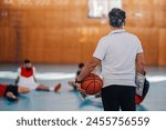 Rear view of an elderly caucasian male basketball coach holding a ball with team in background during a practice and a warmup in a sports hall. Copy space. Background.