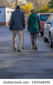 Rear view of an elderly couple walking next to each other and next to parked cars, using a walking stick