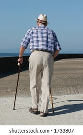 Rear View Of An Elderly Man Walking With Walking Sticks On A Beach Promenade.