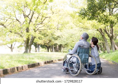 Rear View Of Elderly Man In Wheelchair And Care Helper