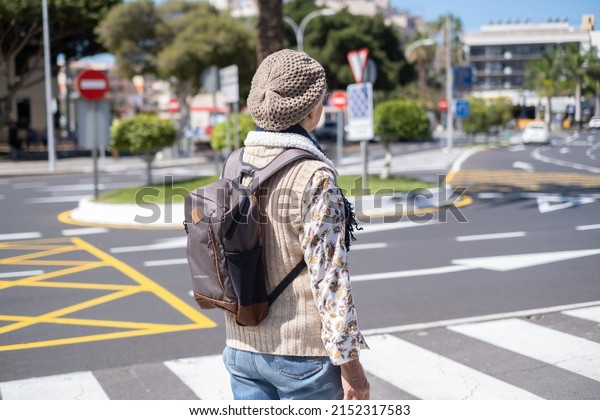 Rear\
view of elderly woman traveler in sunny city center with backpack\
on shoulders while crossing the road on\
crosswalk.