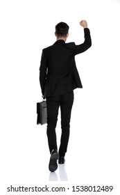 Rear view of a elegant businessman celebrating with his fists in the air and holding a briefcase while wearing a black suit and red tie, moving on white studio background