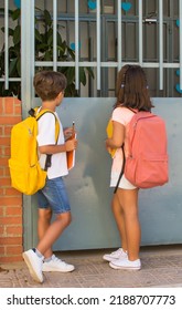 Rear View Of Elementary School Children With School Bags On Their First Day Of School. 2 Children Waiting At The School Gate To Enter.