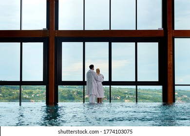 Rear view of embraced couple in bathrobes spending a day at the spa and looking through a window by the swimming pool. Copy space.