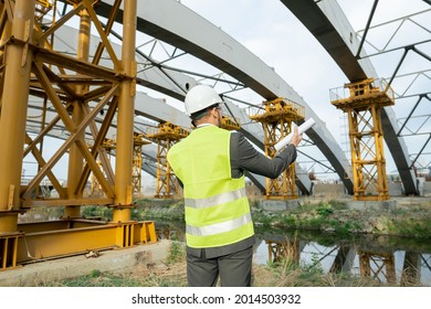 Rear View Of Engineer With Blueprint Looking At Construction Building And Talking On Mobile Phone Outdoors