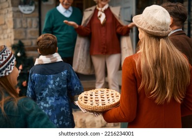 Rear View Of Extended Family Gathering For Thanksgiving. Focus Is On Woman Carrying Sweet Pie.