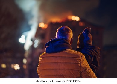 Rear View Of A Father With His Child Watching Their Burning Building. People Watching The Burning Fire Flame With Smoke On The Apartment House Roof. Fire In The City.