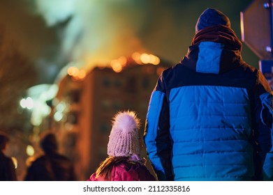 Rear View Of A Father With His Child Watching Their Burning Building. People Watching The Burning Fire Flame With Smoke On The Apartment House Roof. Fire In The City.