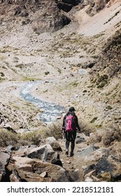 Rear View Of Female Backpacker Hiking Through A Rocky Path Beside A Mountain River. Vertical Shot