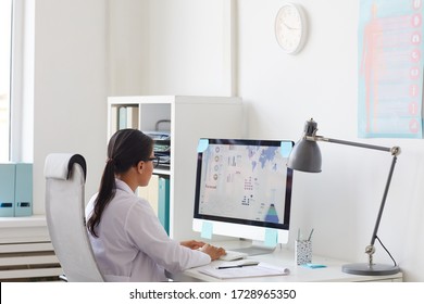 Rear View Of Female Doctor Sitting At The Table In Front Of Computer Monitor And Typing On Computer Keyboard At Office