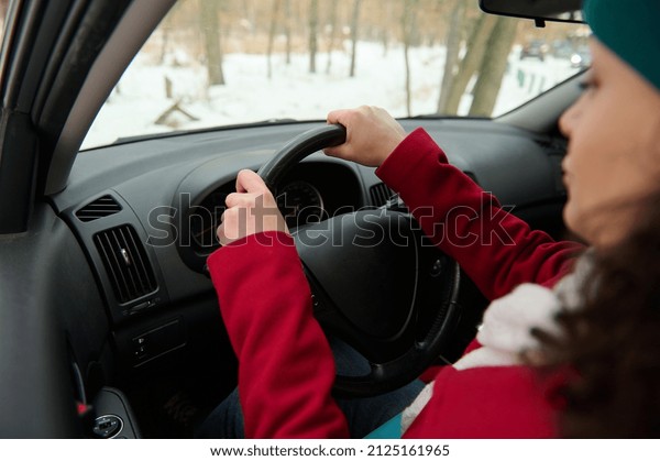 Rear view of a female driver, woman traveler\
holding hands on a steering wheel, sitting on a driver seat of a\
modern car and driving, parking her automobile in a snowy woodland.\
Travel by car concept
