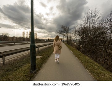 Rear View Of A Female Girl Walking As Pedestrian On A Side Walk Alley Next To Busy Highway Of Cracow Against Dramatic Clouds