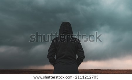 Rear view of female person wearing hooded jacket against dark moody dramatic clouds at sky, woman looking into uncertain ominous future