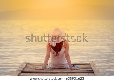 Rear view of female sitting on jetty by the sea at sunset. Summertime concept.   