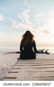 Rear View Of Female Surfer At The Beach Sitting On Jetty With Surfboard At Sunset