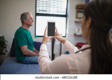 Rear view of female therapist photographing senior male patient from mobile phone against window at hospital ward - Powered by Shutterstock