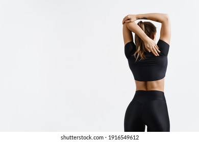 Rear view of fit and sporty fitness woman stratching arms and back before workout exercises. Woman back in black sport clothing standing on white background, training concept.