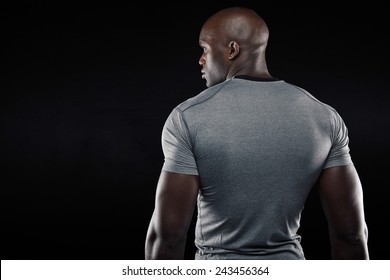 Rear View Of Fit Young Man With Muscular Build Standing Against Black Background. Afro American Fitness Model Looking At Copy Space.