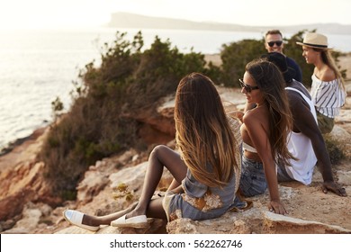 Rear View Of Friends Sitting On Cliff Watching Sunset